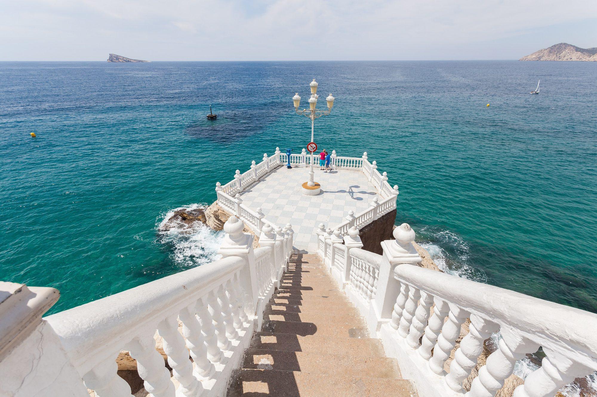 The white balustraded staircase and chequered terrace of the Balcón del Mediterráneo, a lookout on a rocky point above turquoise water, with the Isla de Benidorm on the horizon and a rocky headland at right