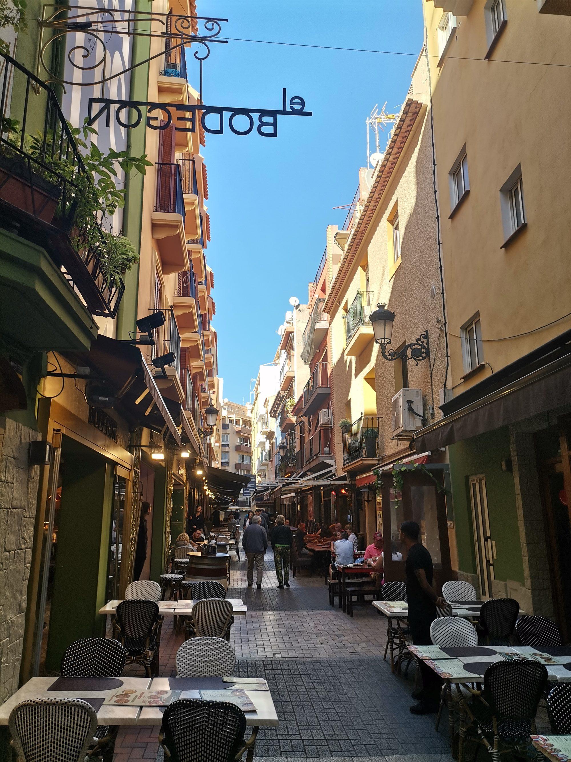 Calle Santo Domingo in Benidorm's old town: a narrow pedestrian lane of restaurant terraces with marble-topped tables and woven bistro chairs, wrought-iron balconies and street lamps, under a strip of blue sky