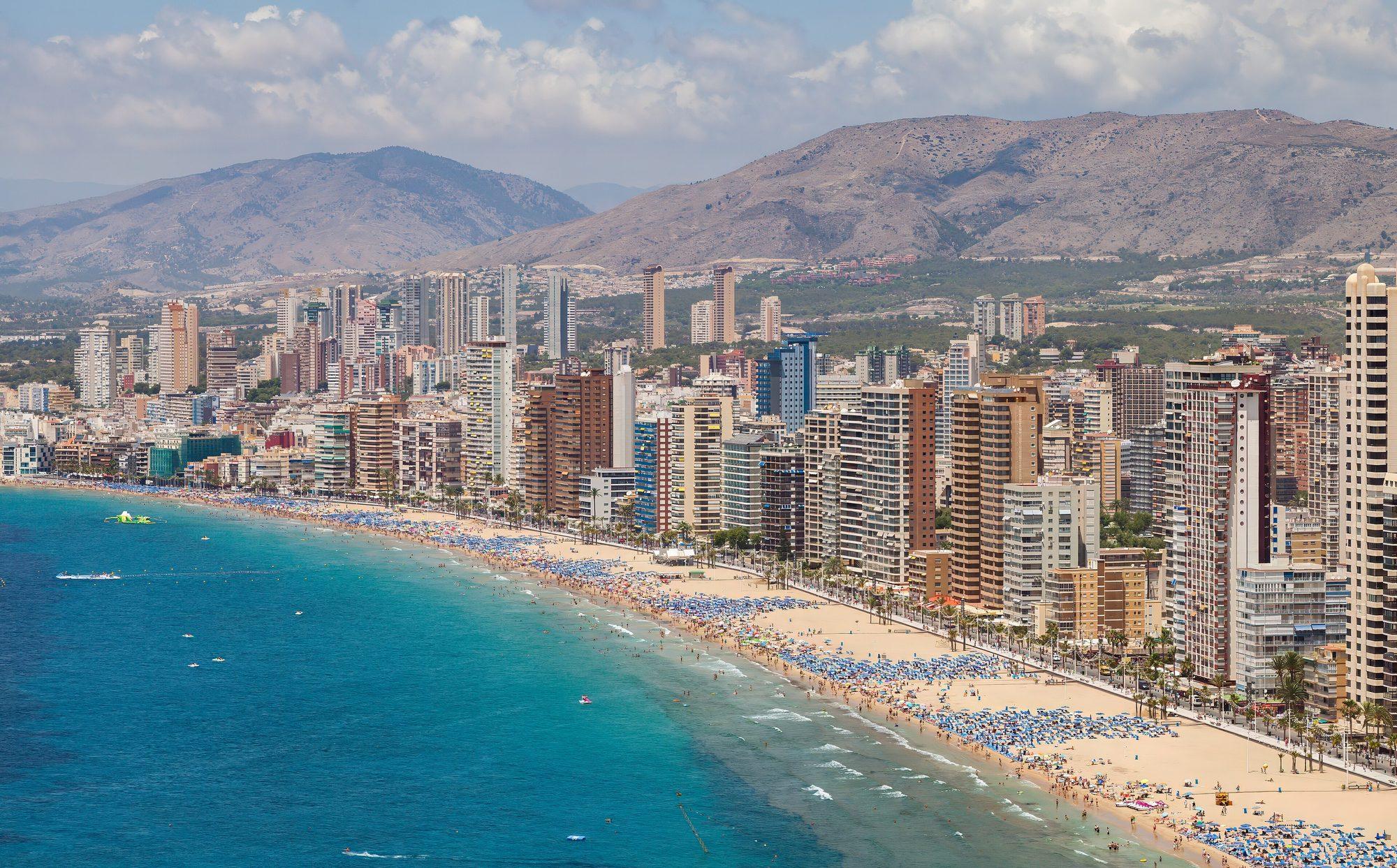 Benidorm's wall of high-rise towers lining the long curve of Playa de Levante, seen from above: turquoise sea, sand packed with blue parasols, and dry mountains behind the city under a partly cloudy sky