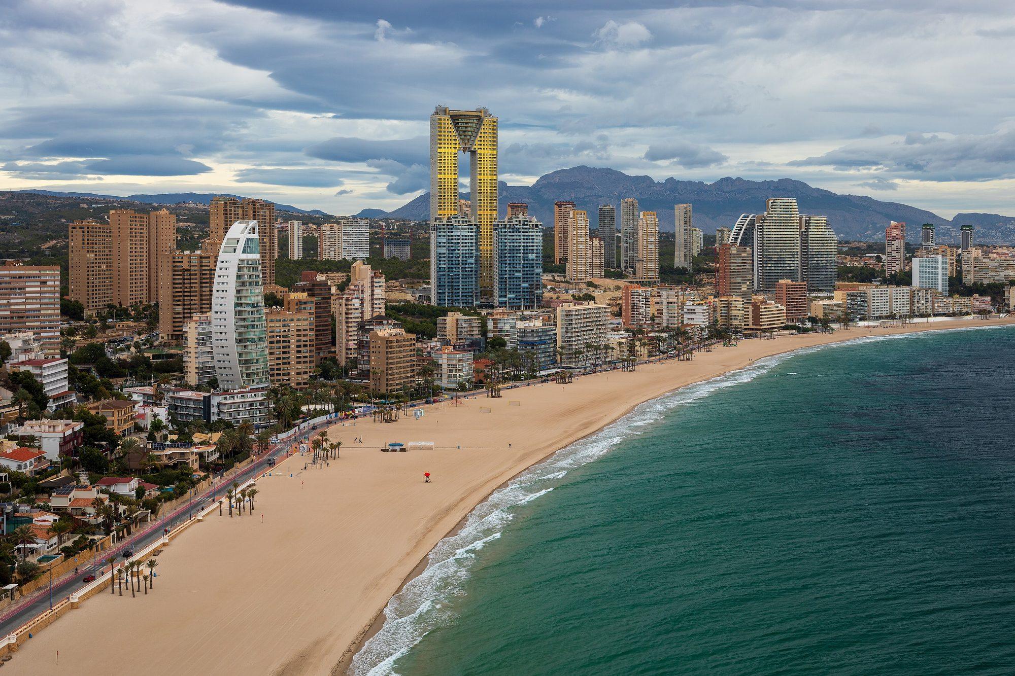 Playa de Poniente seen from above at La Cala: a near-empty curve of sand and surf along the seafront, with the white sail-shaped Delfín Tower, the golden Intempo skyscraper and the city's high-rises rising against the mountains under a cloudy sky