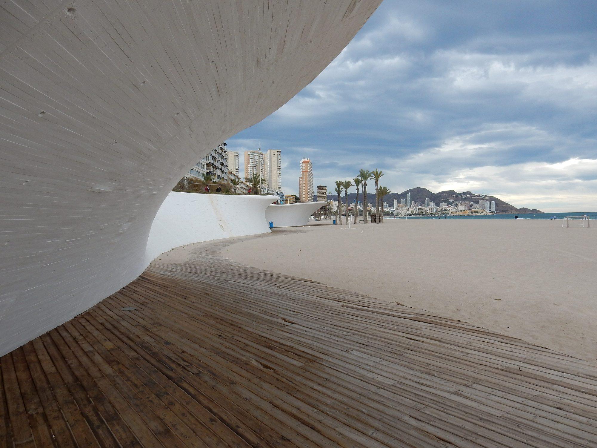 The sculpted white concrete waves of the Paseo de Poniente promenade curving over a timber boardwalk on Playa de Poniente, with palm trees, apartment towers and hills beyond under a cloudy sky