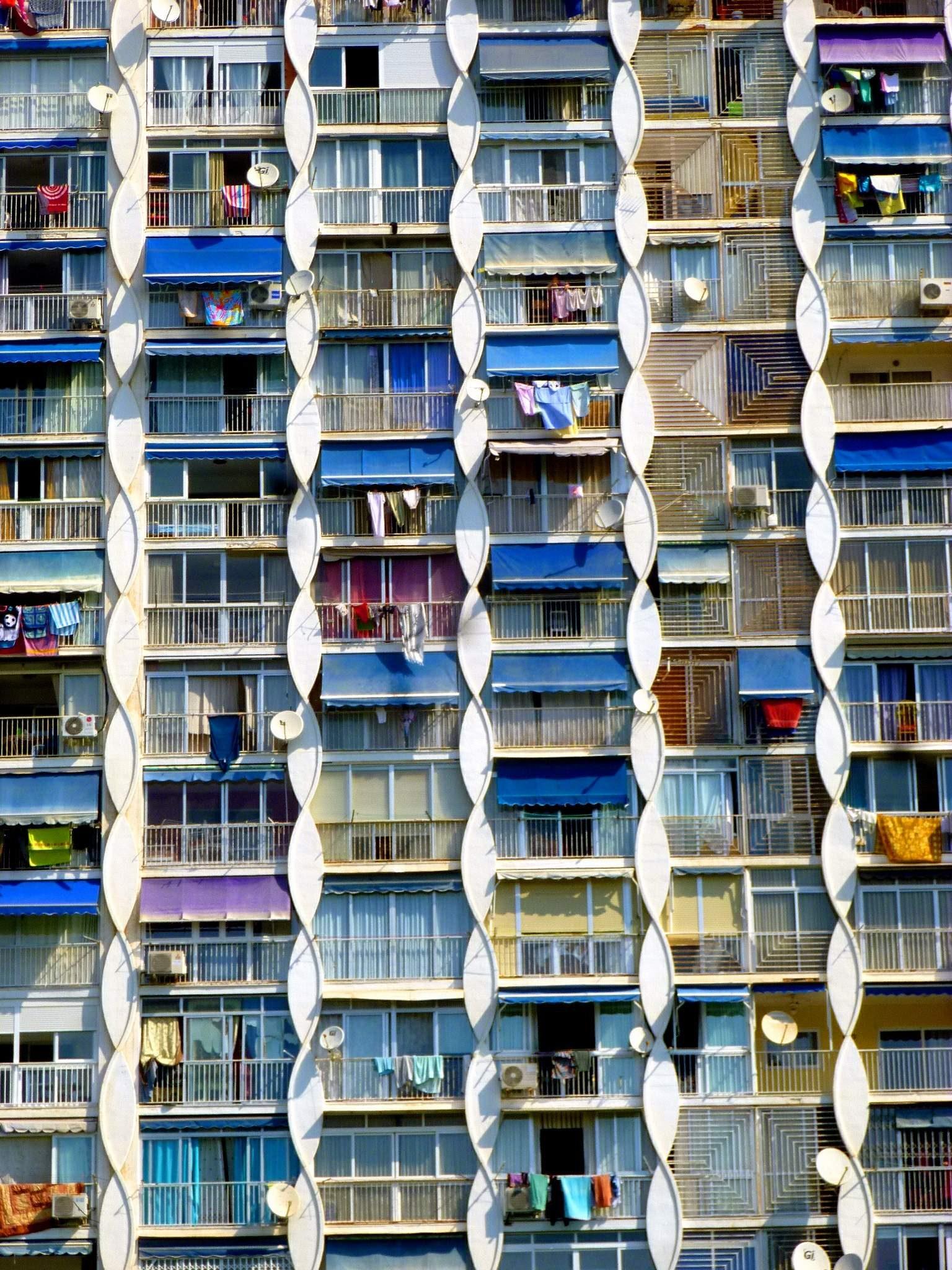 Close-up of the façade of the Playmon Fiesta apartment tower: stacked balconies with blue and purple awnings, satellite dishes and drying laundry, framed by white wavy concrete fins